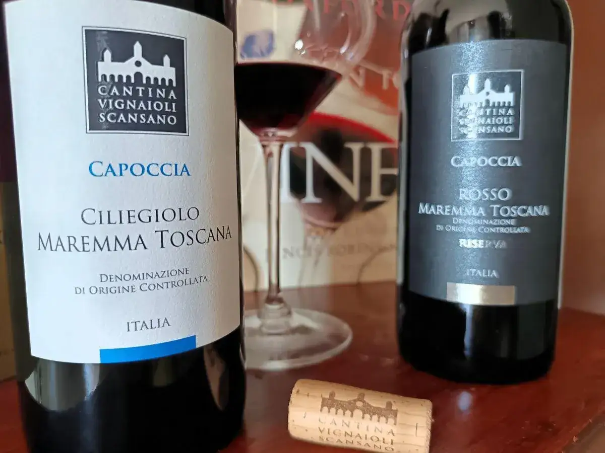 Due bottiglie di vino rosso della Cantina Vignaioli Scansano, una Ciliegiolo Maremma Toscana e una Riserva. Un tappo di sughero e un calice di vino completano la scena. Scansano, cosa vedere per gli amanti del buon vino.