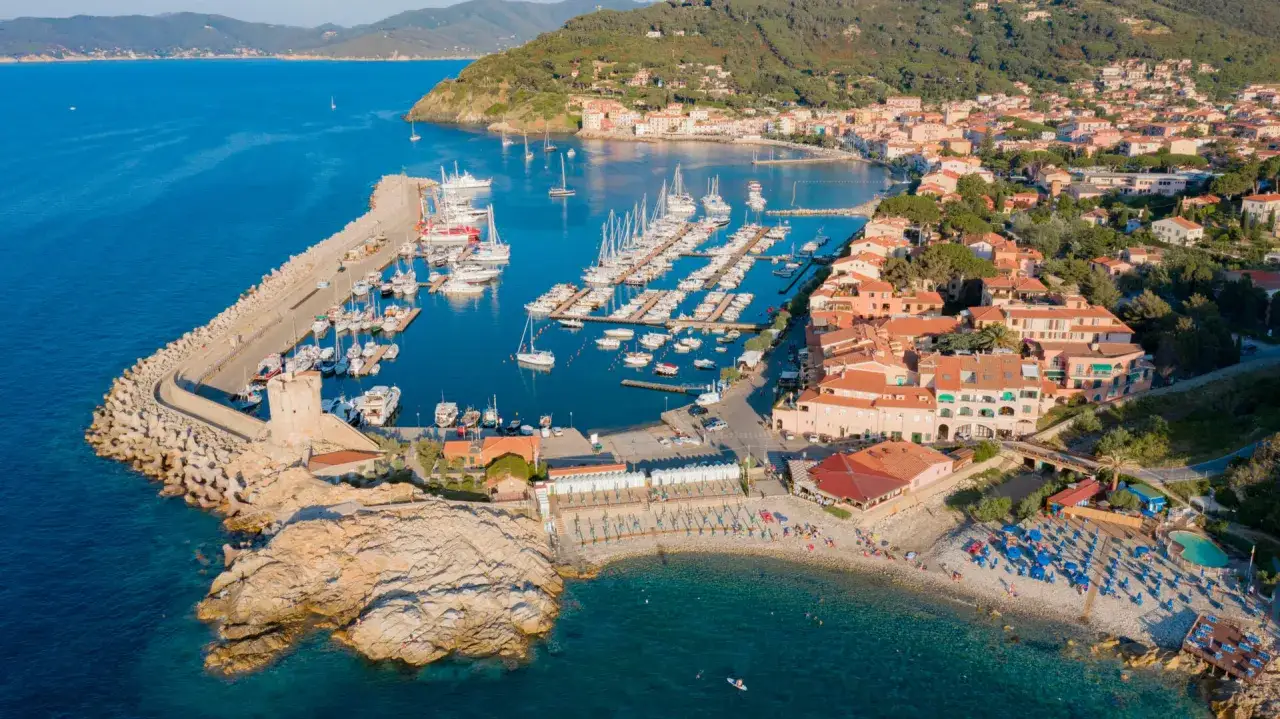 Vista aerea del porto turistico e della spiaggia di Porto Azzurro, Isola d'Elba, con case colorate e un'antica torre.