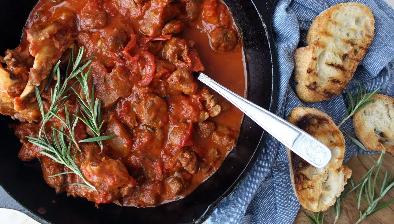 Un saporito buglione di agnello in padella, guarnito con rosmarino fresco, accompagnato da fette di pane tostato.