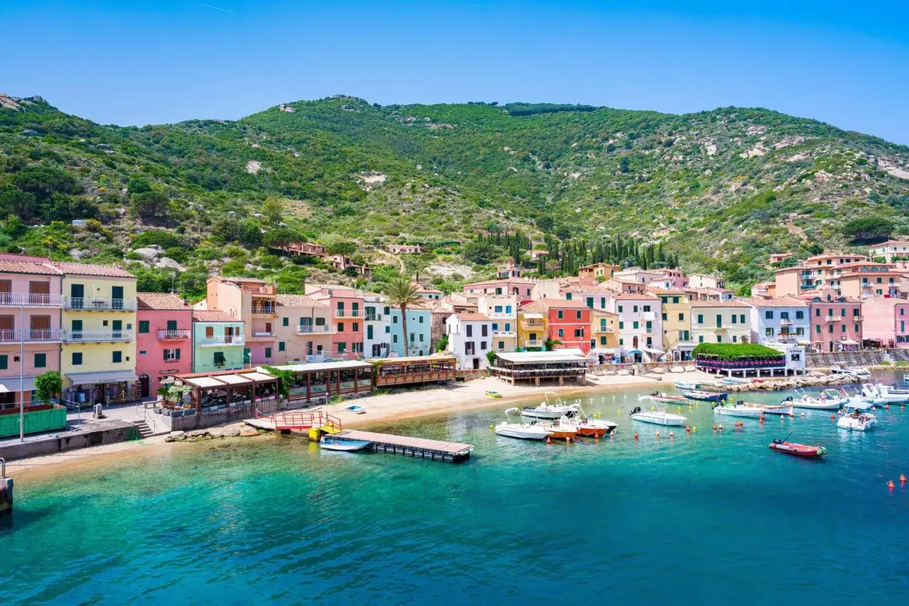 Case colorate affacciate sul mare cristallino di Porto Santo Stefano, con barche ormeggiate e una collina verdeggiante sullo sfondo.