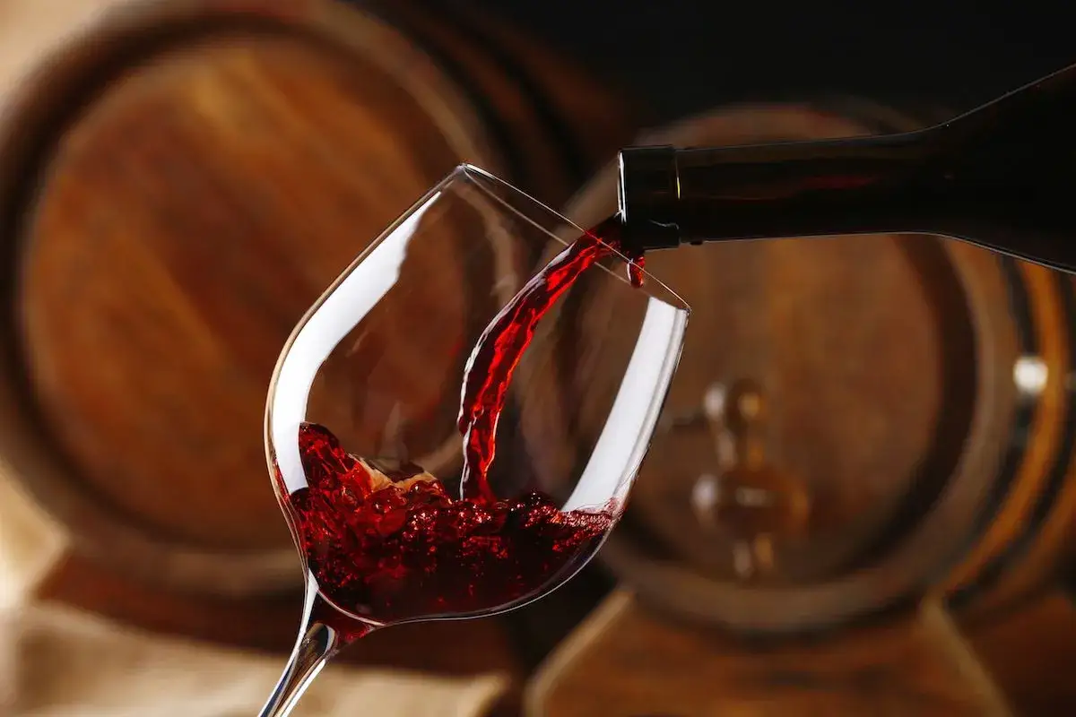 Versando vino rosso corposo, dal gusto tannico, in un calice, con botti di legno sullo sfondo.
