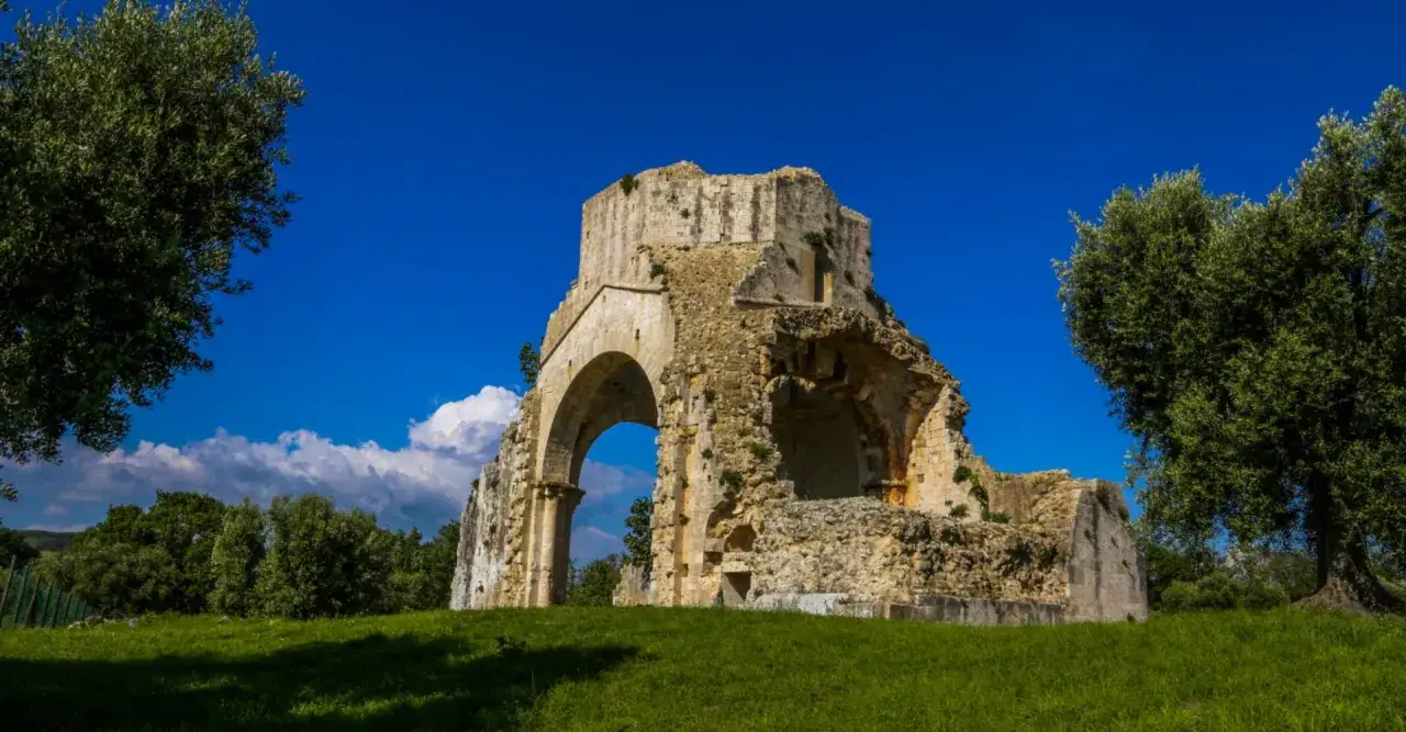 Rovine dell'abbazia di San Bruzio sotto un cielo azzurro, con alberi verdi e nuvole bianche.