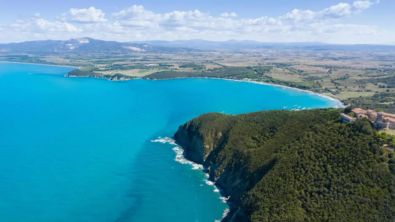 Costa toscana con mare azzurro e vegetazione lussureggiante. Si intravede un borgo storico, forse dove si trova Castagneto Carducci.