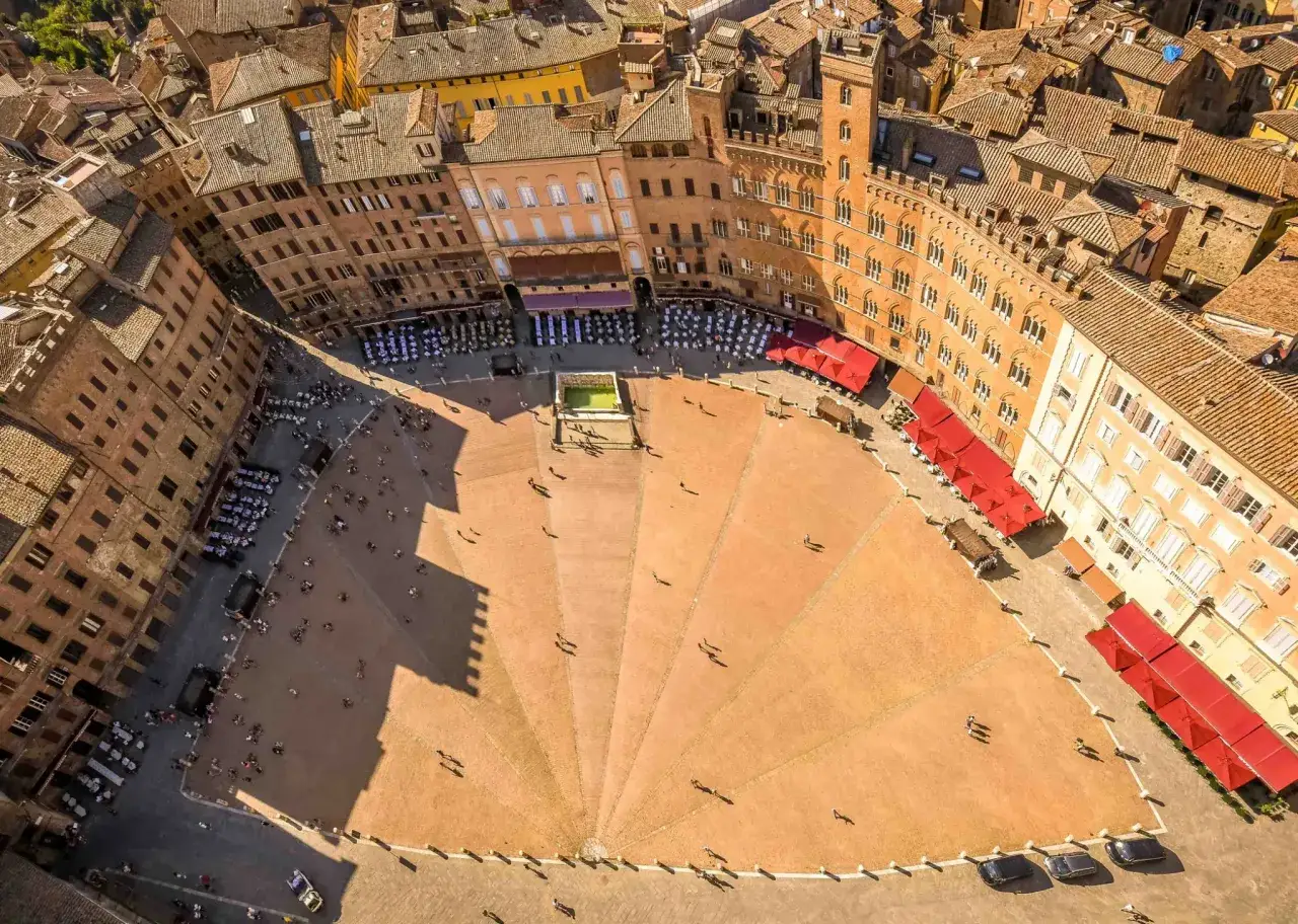 Vista aerea della Piazza del Campo a Siena, con la sua caratteristica forma a conchiglia, circondata da palazzi storici e animata da persone.