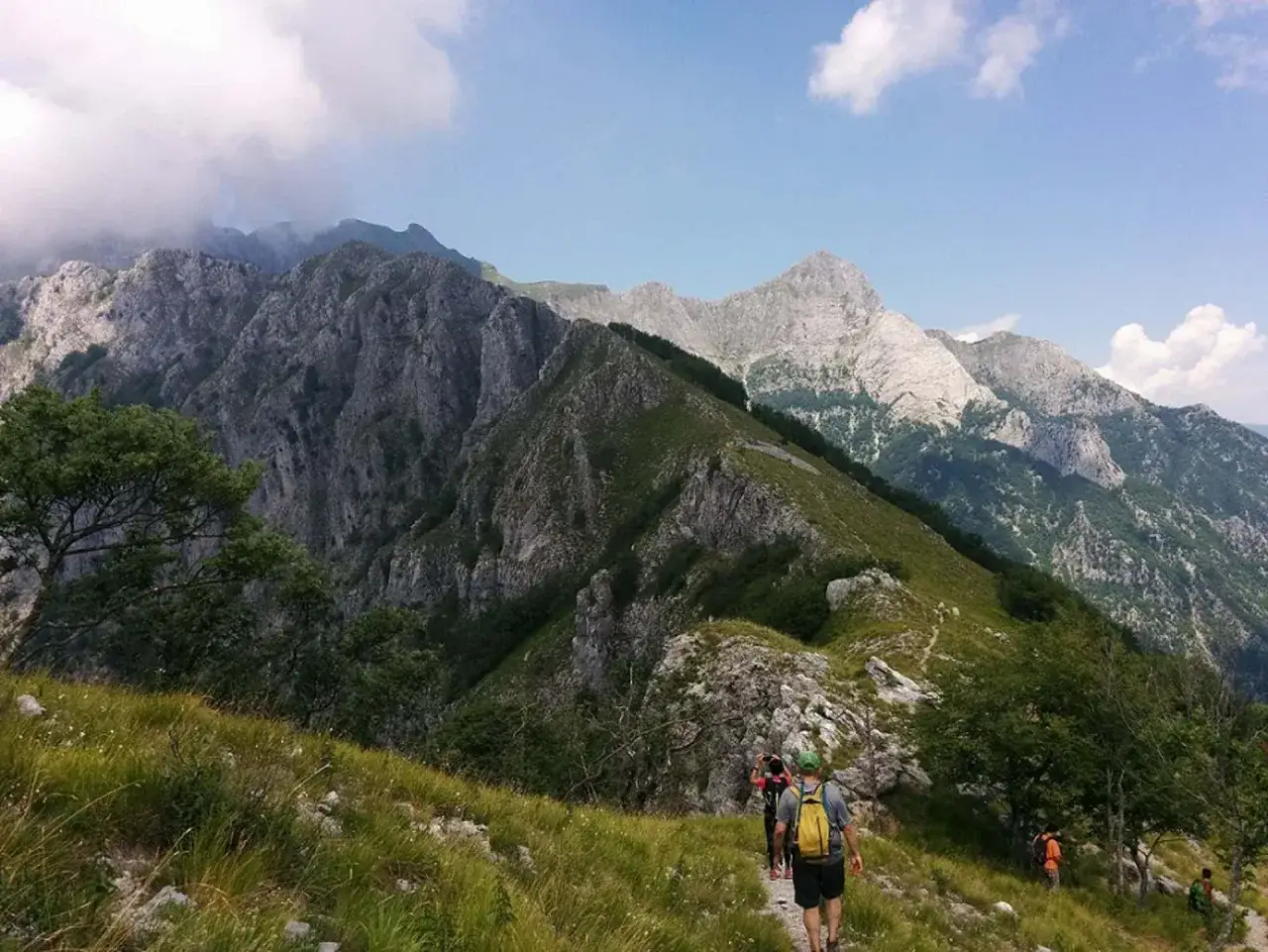 Sentiero di trekking nelle Alpi Apuane, con escursionisti che camminano su un crinale erboso tra montagne rocciose e cielo azzurro.