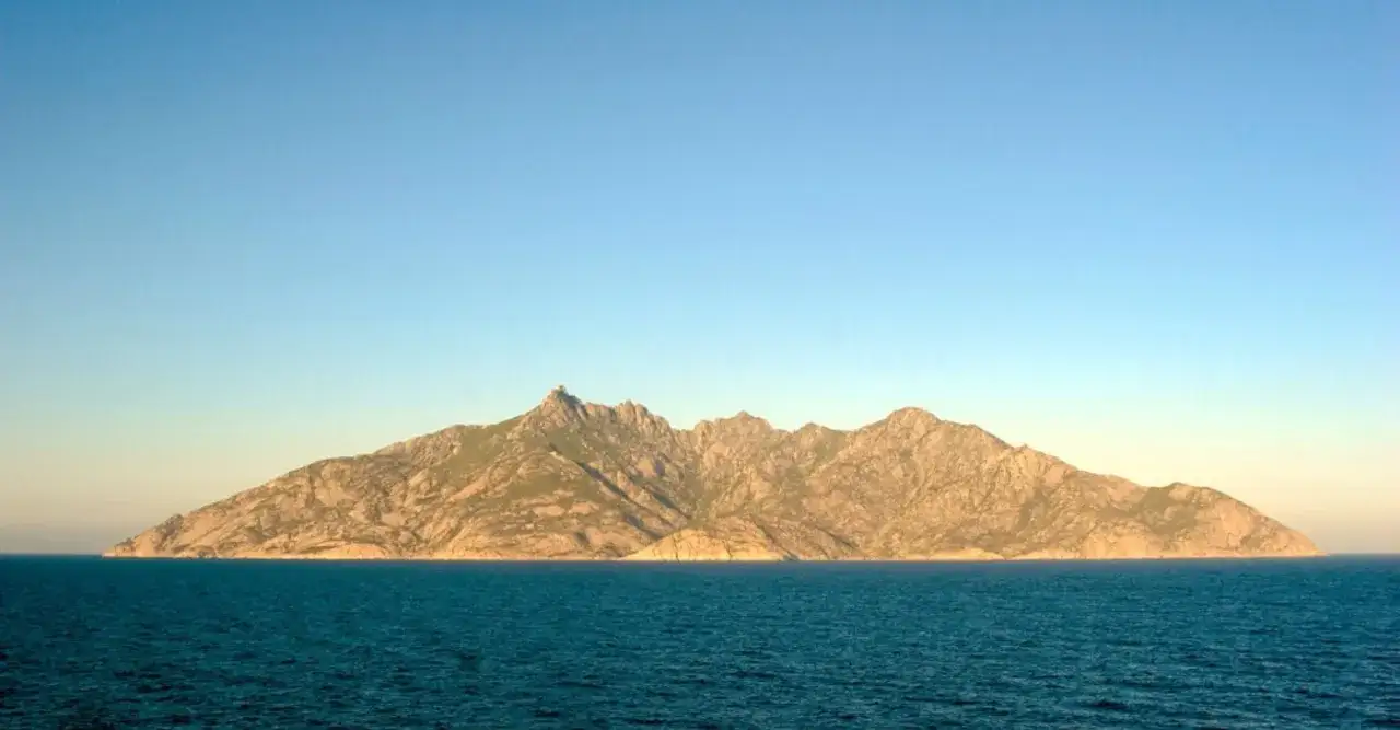 Isola selvaggia nel mare, parte dei parchi della Toscana, con montagne rocciose e vegetazione bassa sotto un cielo azzurro.