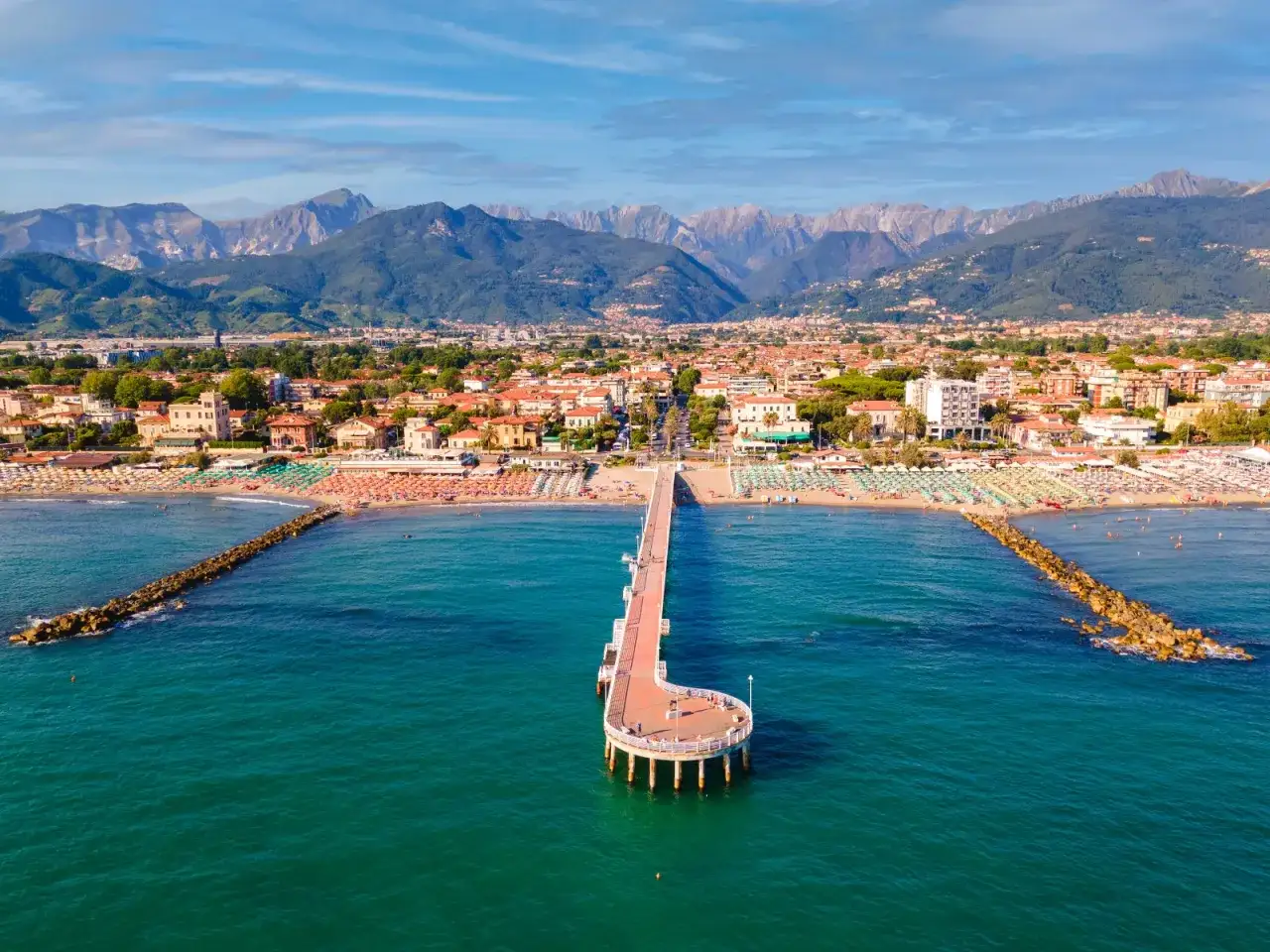 Un lungo molo si estende nel mare turchese di Marina di Massa, con montagne sullo sfondo. Le spiagge sono piene di ombrelloni colorati.