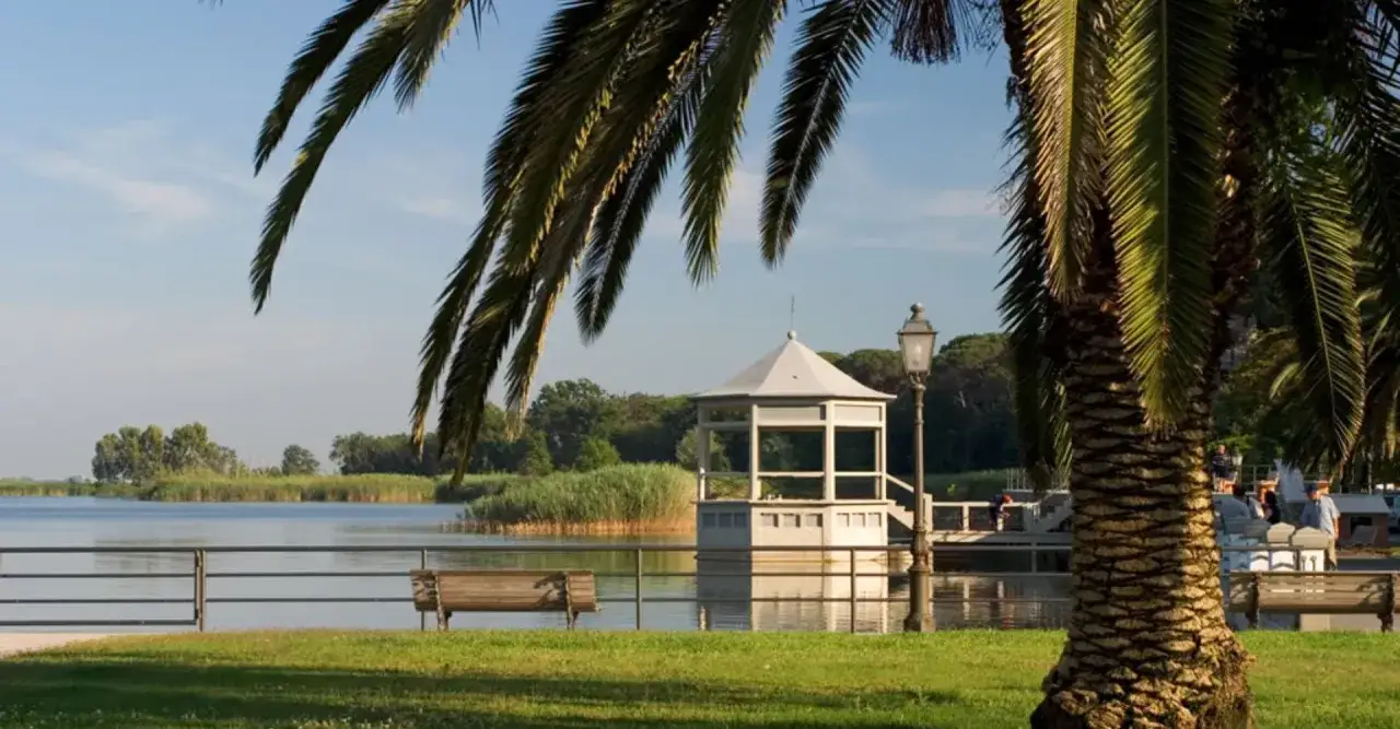 Un gazebo bianco si affaccia sul lago, vicino a una torre, nel centro storico. Palme e panchine completano la scena.