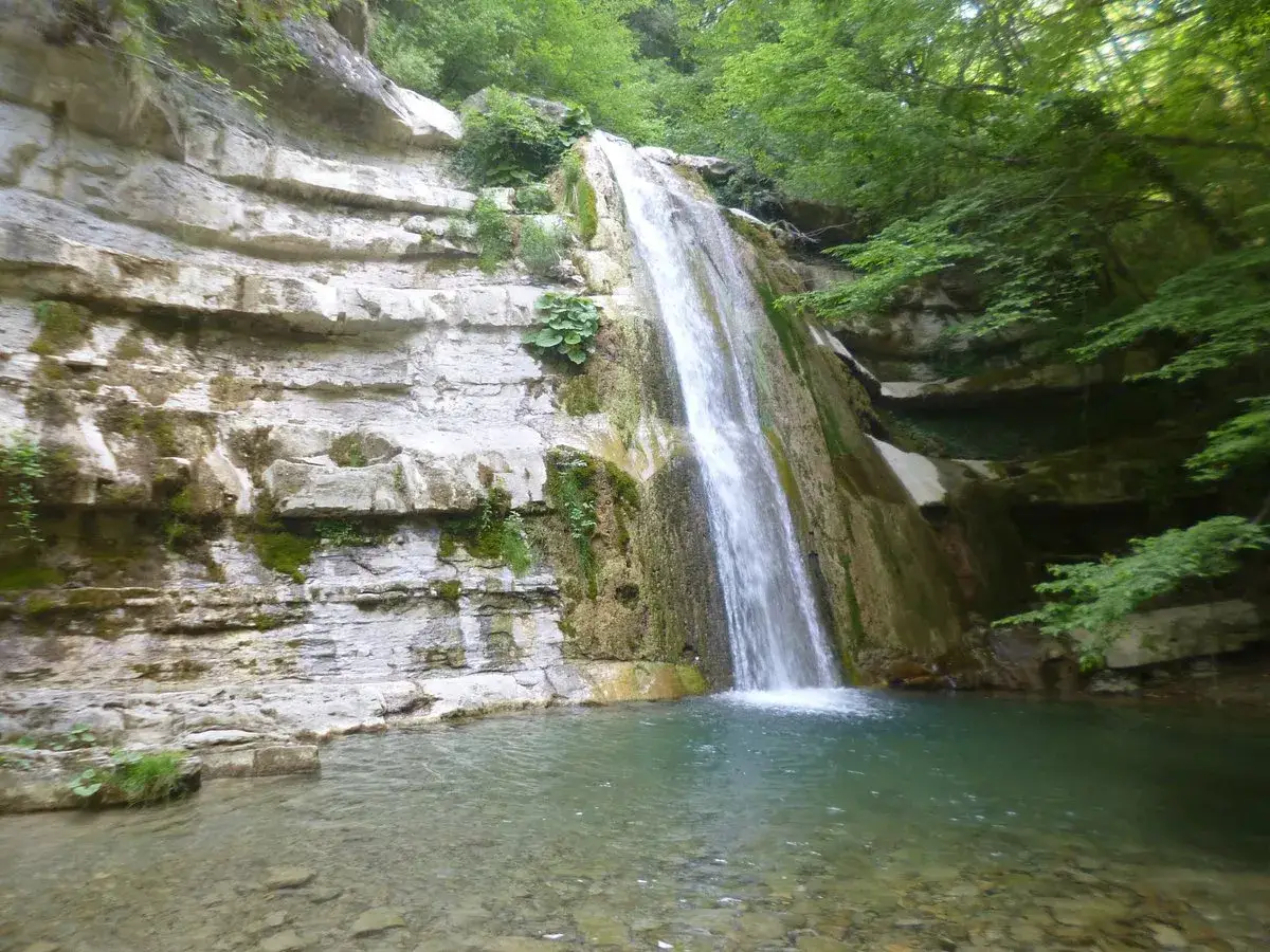 Cascata fragorosa che si getta in un laghetto cristallino, circondata da rocce stratificate e vegetazione lussureggiante. L'acqua cheta del laghetto riflette il verde intenso del bosco.