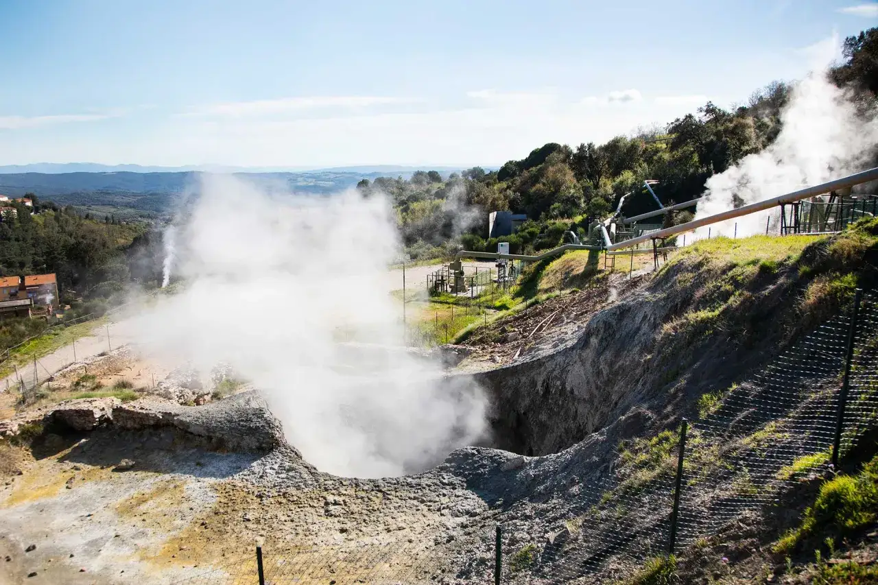 Un geyser in Italia, con vapore che sale da una voragine e tubature industriali sullo sfondo.