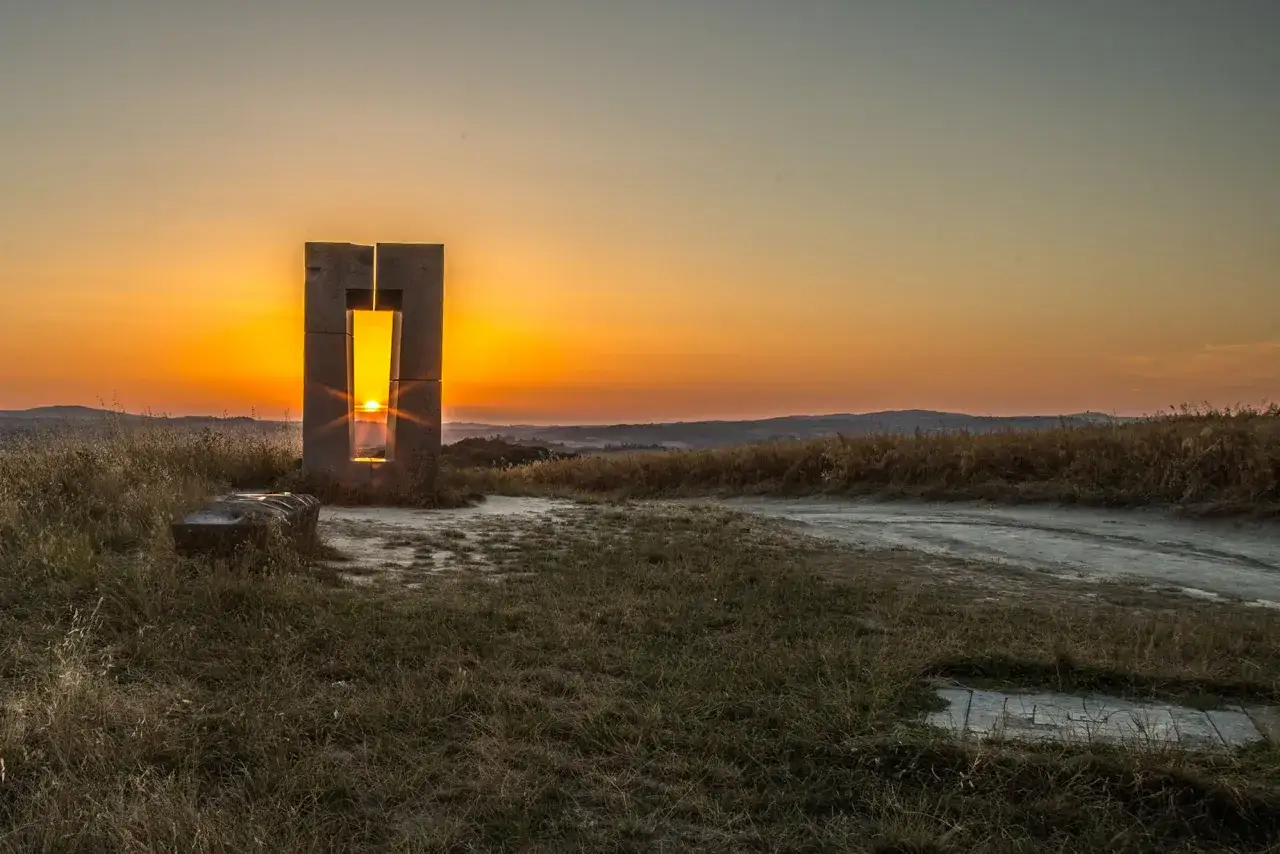 Tramonto dorato attraverso il sito transitorio di Asciano, con un monumento in pietra che incornicia il sole e un paesaggio collinare sullo sfondo.