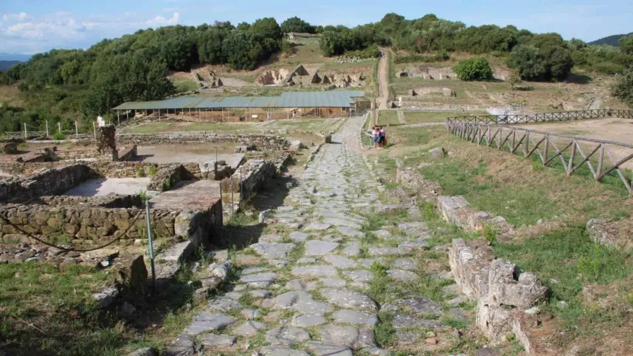 Antica strada romana conduce alle rovine di Vetulonia, con resti di edifici e un tetto protettivo. Le terme di Vetulonia emergono dal paesaggio.