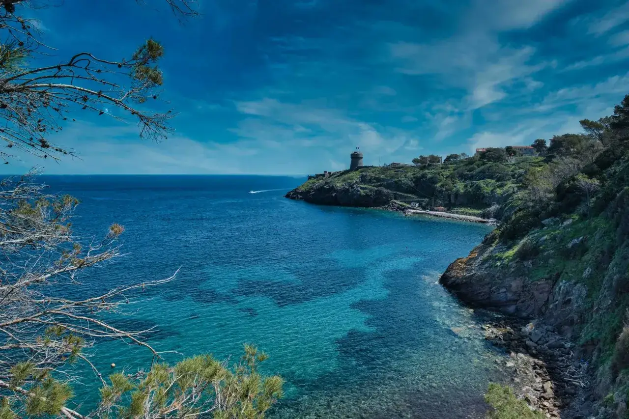 Vista mozzafiato su una baia cristallina di Capraia isola, con un faro in lontananza e vegetazione mediterranea.