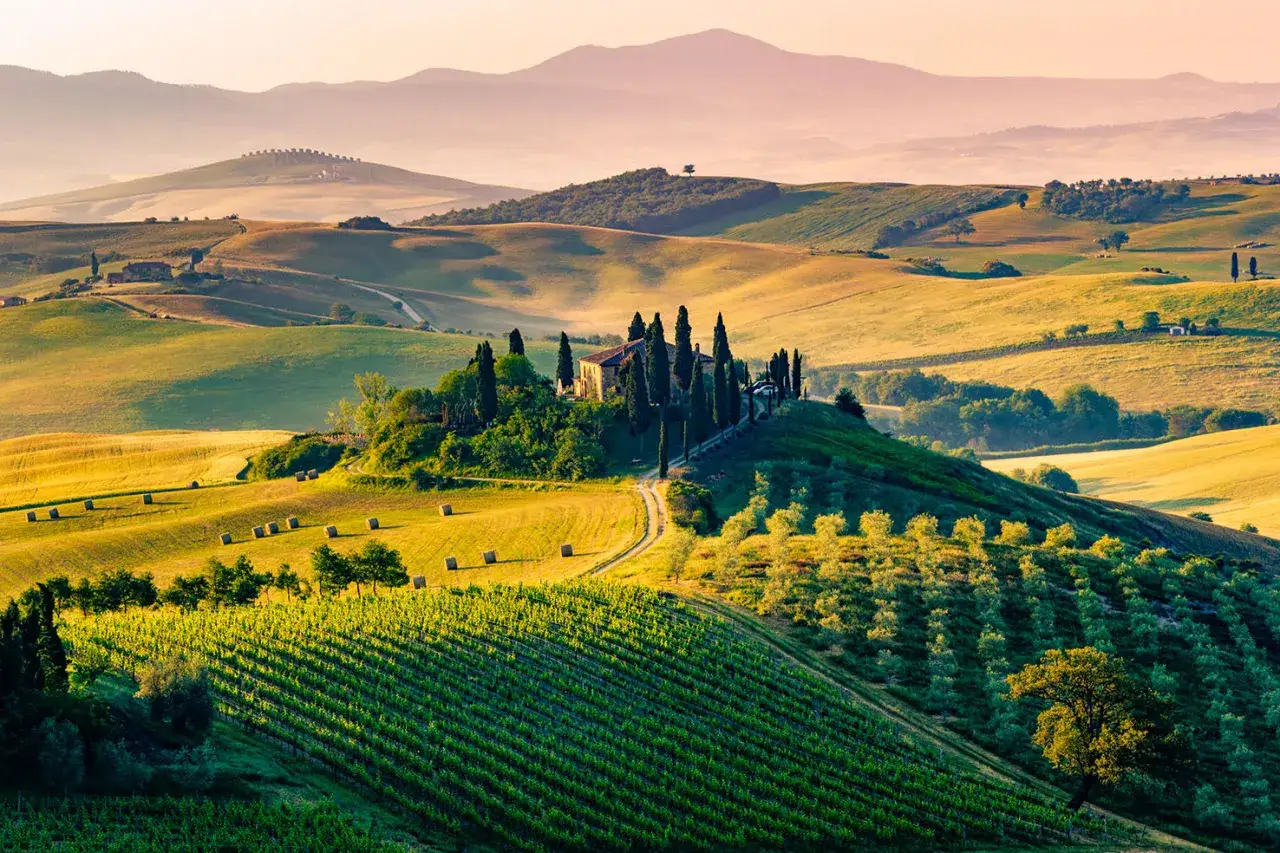 Dolci colline dell'entroterra toscano al tramonto, con una villa su una collina, filari di vite e balle di fieno.