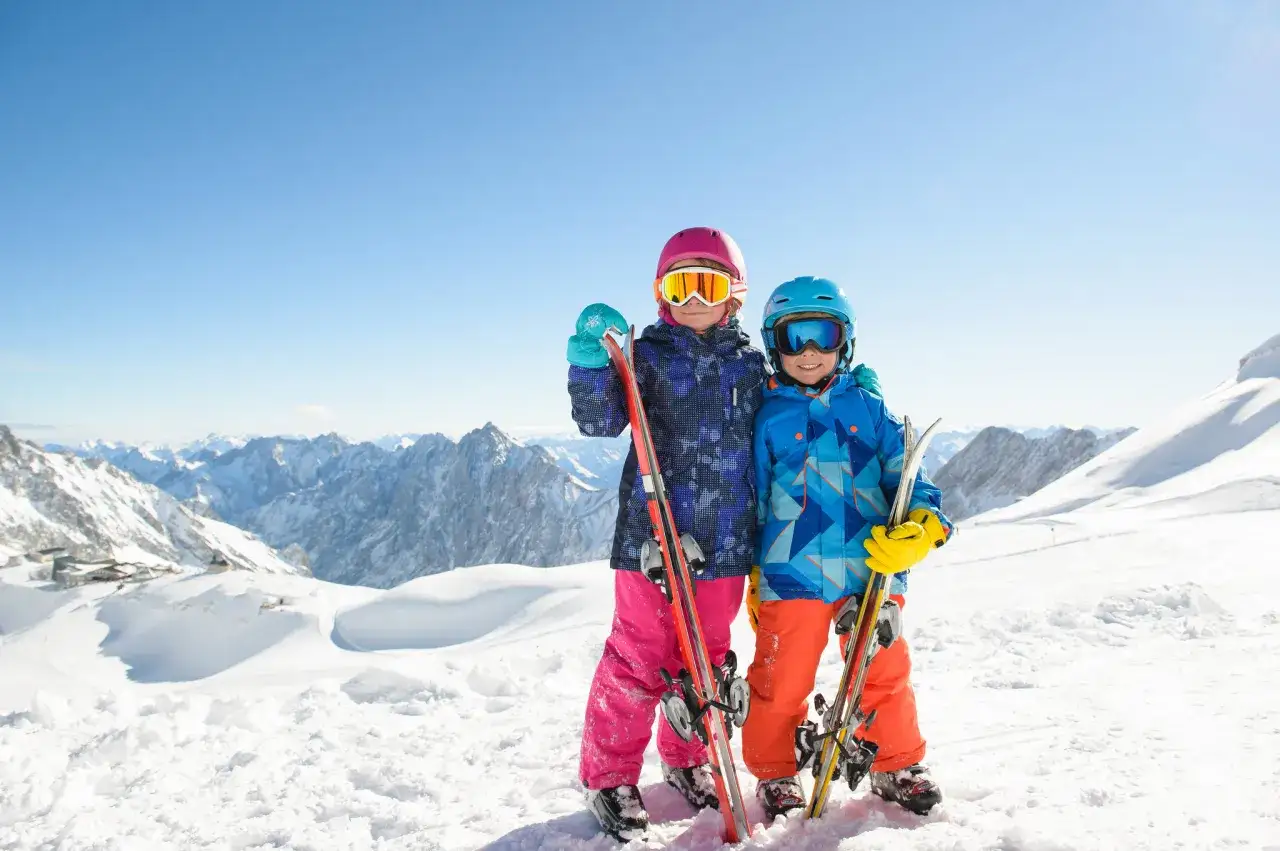 Due bambini con sci e abbigliamento da neve sorridono in cima a una montagna innevata, con un panorama mozzafiato. Sembra il centro di Abetone in una giornata perfetta.