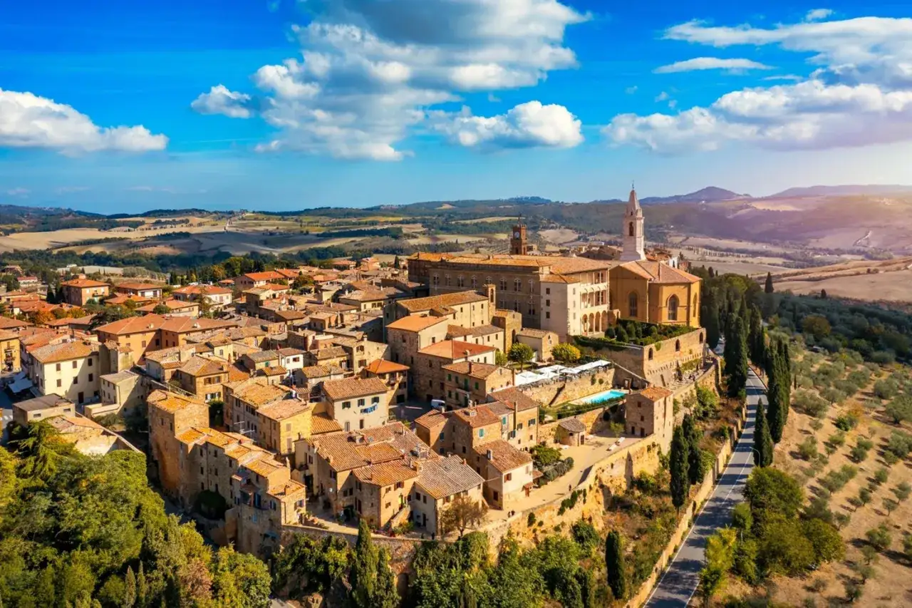 Panoramica aerea di un borgo medievale toscano, forse Montalcino o Montepulciano, con tetti in cotto e una chiesa imponente.